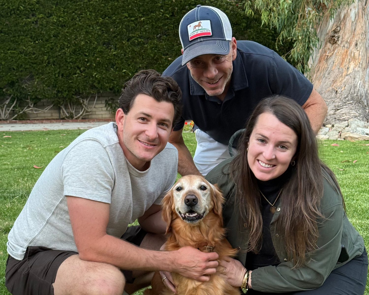 Three people crouching on a green lawn with a golden retriever dog in the center, outdoors with trees and shrubs in the background.